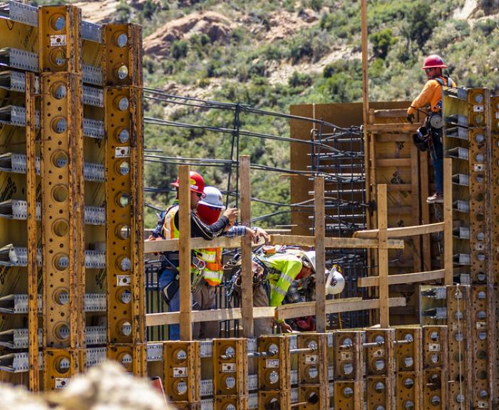 Construction workers in safety gear working on a bridge framework with metal supports and green hills behind.