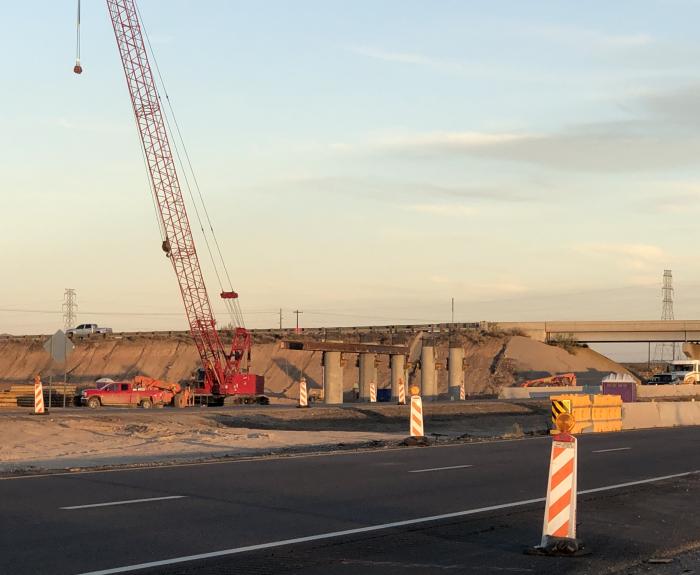 A construction site with a red crane, concrete pillars, sand piles, and caution barriers near a highway under an evening sky.