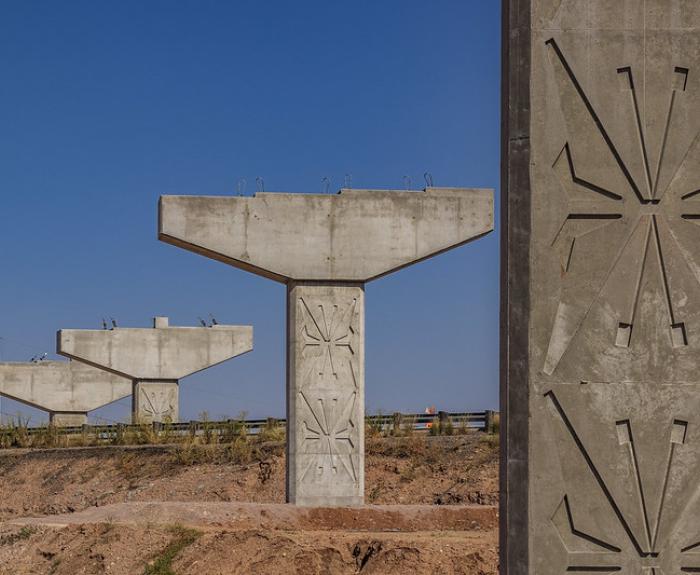 Three unfinished concrete bridge supports stand on a dirt embankment under a clear blue sky, with stylized monarch butterfly patterns visible on the closest pillar.