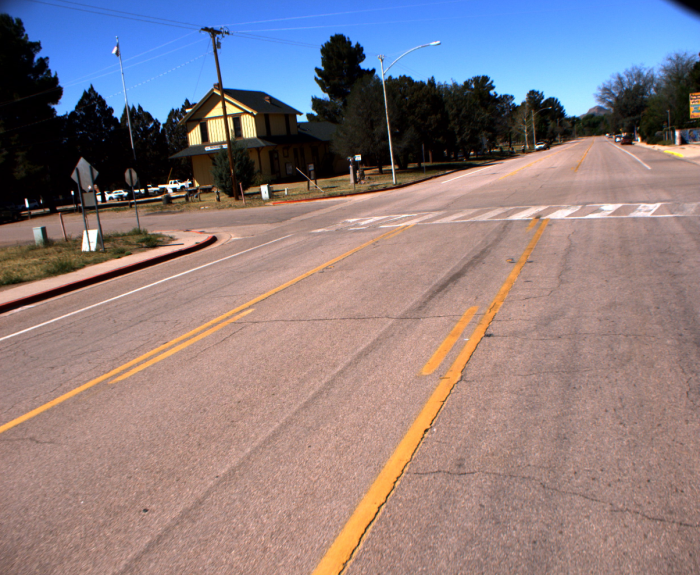 A quiet, empty street with yellow lines and a train depot surrounded by trees under a clear blue sky.