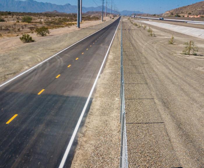 A paved road runs parallel to a canal in a dry, desert landscape with mountains in the distance.