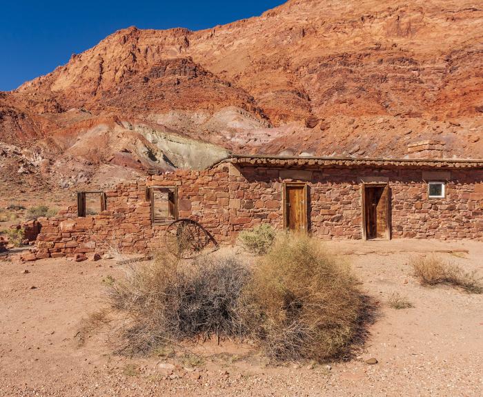 A small, abandoned stone building with wooden doors stands in a dry desert landscape against a backdrop of red rocky hills under a clear blue sky.