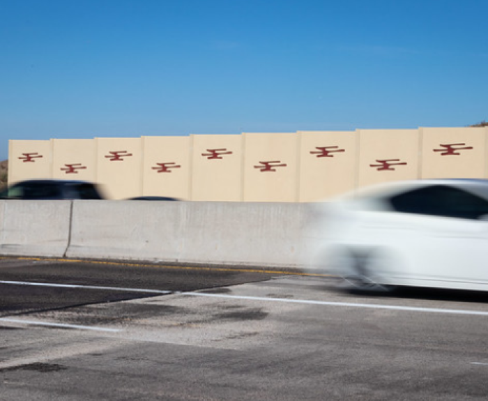 A white car speeds past a beige highway sound wall decorated with red geometric designs.
