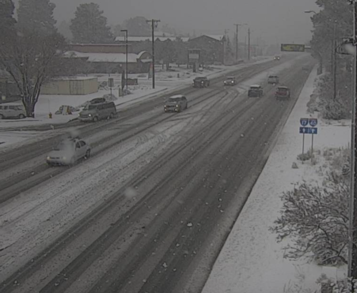 Several cars drive on a snow-covered road during snowfall, with light snow accumulation visible on the ground, trees, and buildings.
