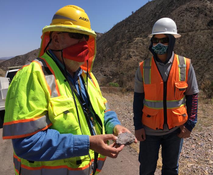Two workers in safety vests and helmets, one holding a rock sample, stand on a roadside with rocky hills in the background. Both are wearing face masks.
