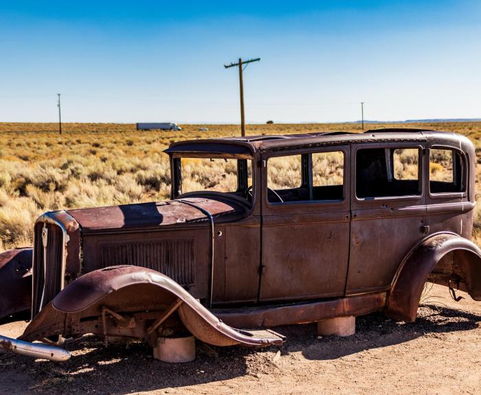 A rusted, vintage car sits abandoned in a dry, grassy landscape under a clear blue sky.