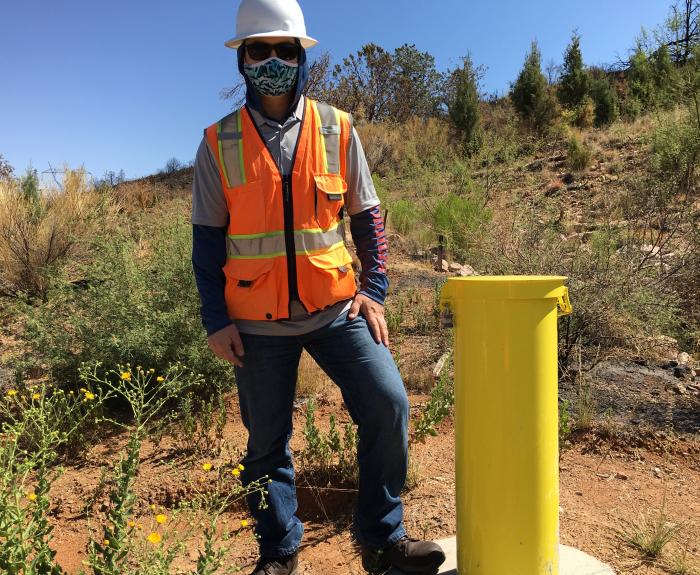 Person wearing a hard hat, safety vest, mask, and sunglasses stands outdoors next to a yellow post in a dry, brushy area.