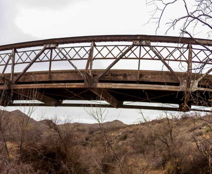 A weathered metal truss bridge the Leslie Creek Bridge spans a dry, brush-filled ravine with barren trees and distant hills under a cloudy sky.