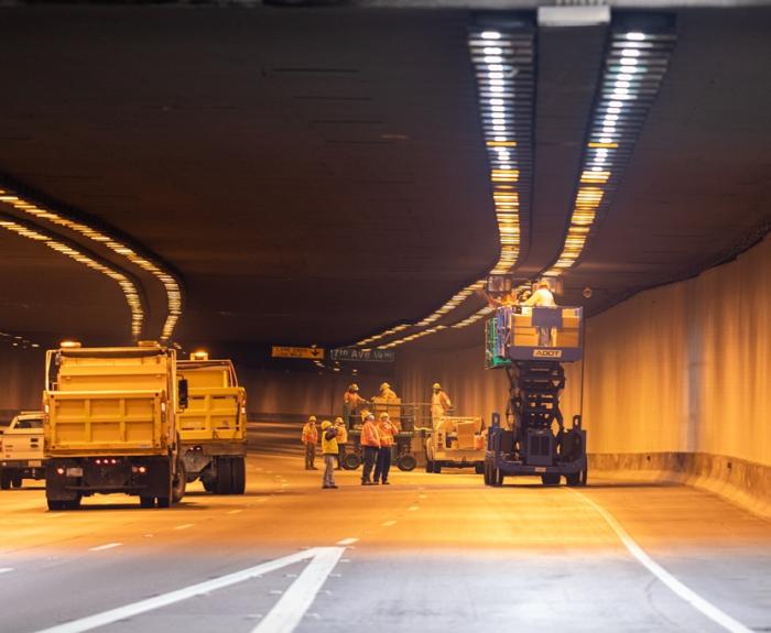 Workers in safety gear perform maintenance in a tunnel using a lift and yellow trucks, with traffic lanes closed and overhead lights illuminated.