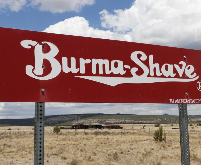 Red Burma-Shave sign stands in a dry, open landscape under a partly cloudy sky.