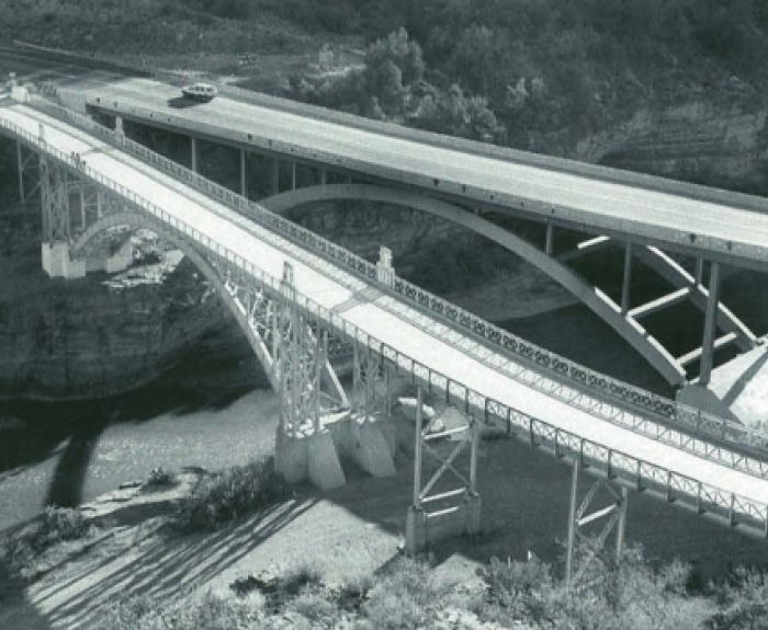 Two parallel bridges span a river, one modern with smooth arches, the other older with intricate truss design.