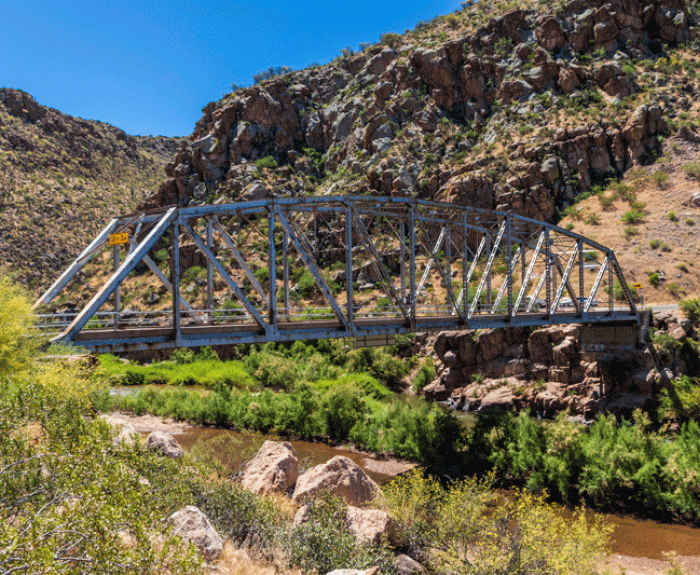 Steel truss bridge spans a rocky canyon with green shrubs and a river below, under a clear blue sky.