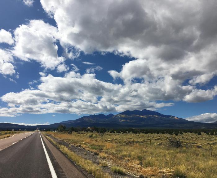A highway runs toward distant mountains under a blue sky filled with large, fluffy clouds.