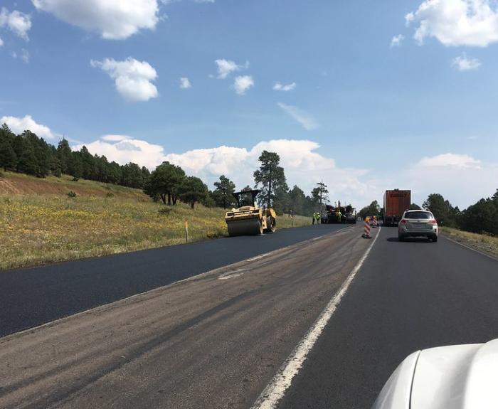 Road construction with vehicles and workers paving a rural highway under a partly cloudy sky.