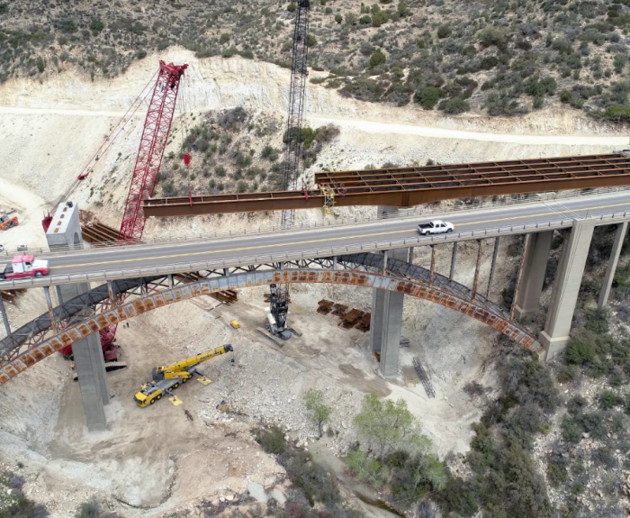 Aerial view of the Pinto Creek Bridge in Arizona under construction with cranes, trucks, and equipment in a hilly area.