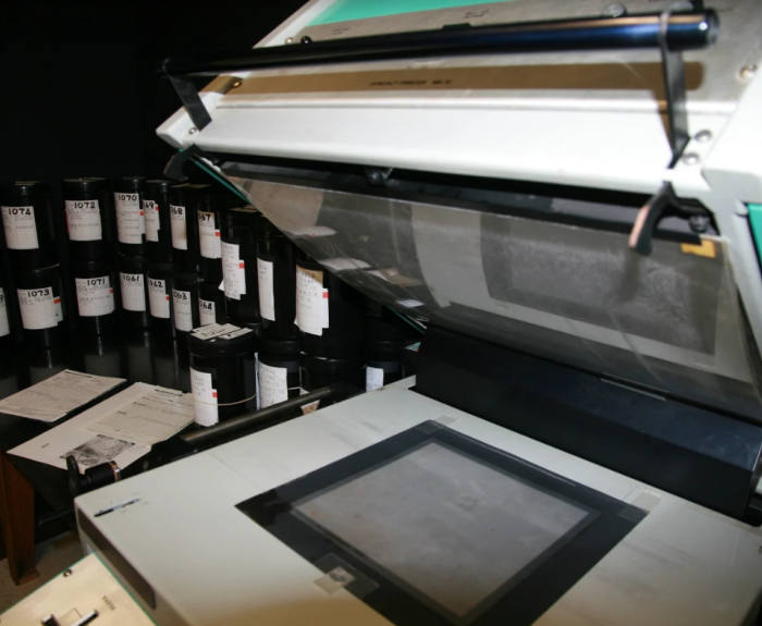 A microfilm reader with reels of microfilm and papers on a nearby table in a dimly lit room.