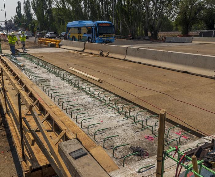 Construction workers build the Rio de Flag Bridge in Flagstaff Arizona. A public transit bus and traffic barriers are visible in the background.