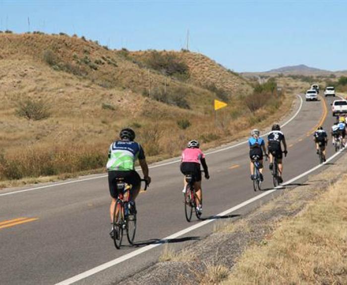 Several cyclists ride uphill on a two-lane rural road with cars following behind. The landscape is dry with grassy hills on both sides.
