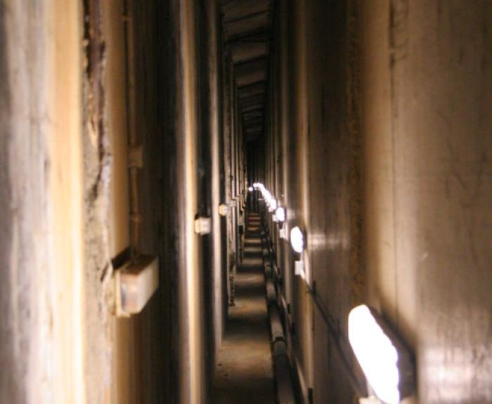 A narrow, dimly lit concrete corridor with exposed pipes and wall-mounted lights running along its length.