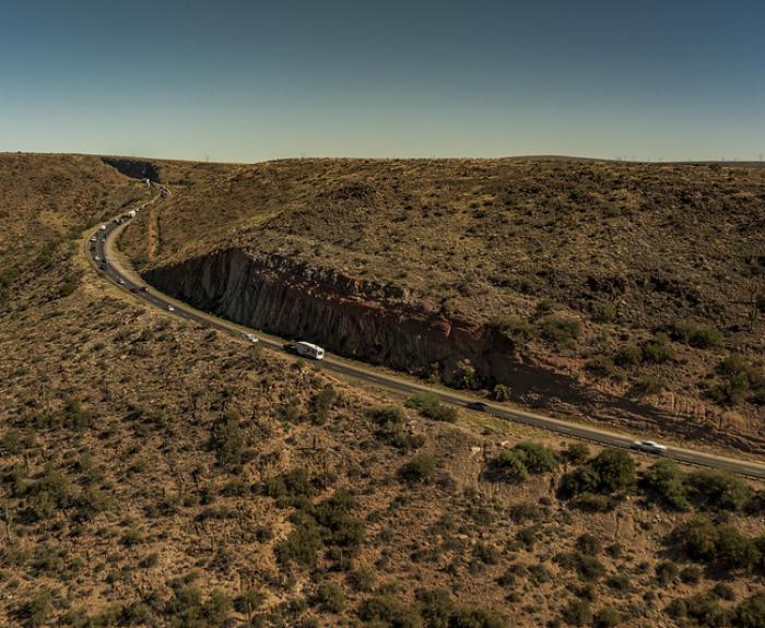 A winding road with cars cuts through a dry, hilly landscape under a clear blue sky.