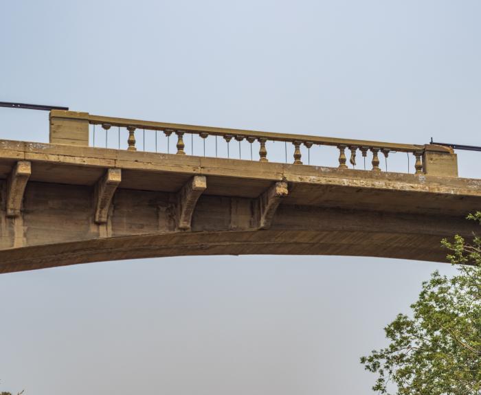 An old concrete bridge with visible railings and support beams, photographed from below against a clear sky with tree branches in the foreground.