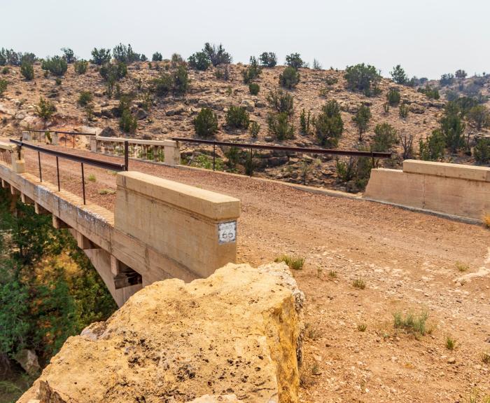 A narrow, weathered concrete bridge with metal railings crosses a dry, rocky landscape with sparse trees and shrubs.