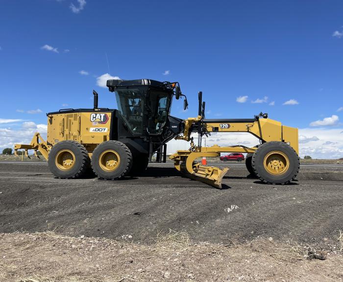 A yellow Caterpillar road grader working on a dirt road under a blue sky with scattered clouds.