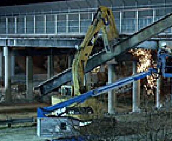A construction worker uses a lift to cut a large metal beam with sparks flying, while an excavator supports the beam near a bridge at night.