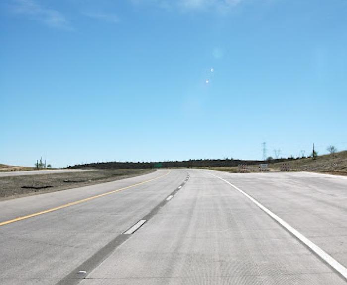 A wide, empty concrete highway curves gently under a clear blue sky with wispy clouds and sunlight glare near the horizon.