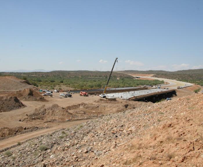 A construction site with vehicles, dirt piles, and a crane building a bridge over a road in a dry, hilly landscape under a clear sky.