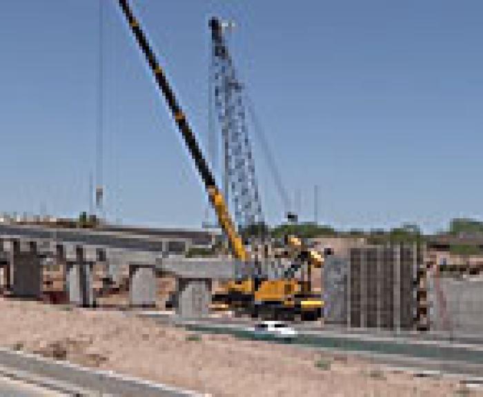 A crane lifts a large concrete section during bridge construction over a highway, with several support columns and a car driving below.