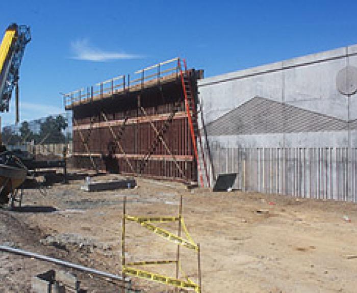 A construction site with a crane, scaffolding, caution tape, dirt ground, and a large unfinished concrete wall under a clear blue sky.
