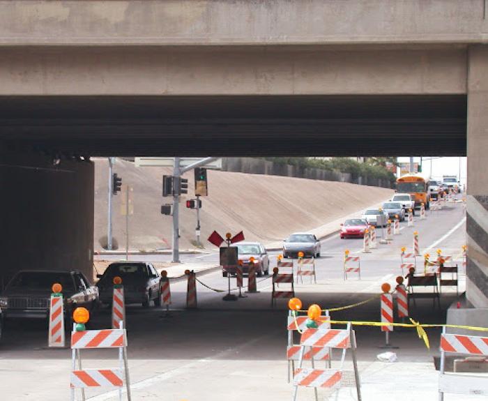 Cars detour under a bridge with road construction signs and barriers directing traffic.