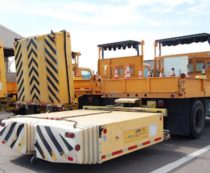 Yellow highway maintenance trucks with crash attenuators parked in a lot, safety cones on truck beds.