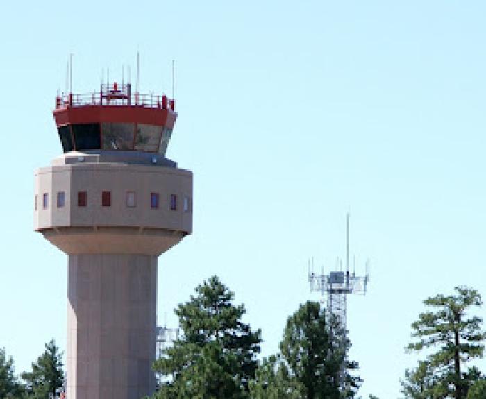 An air traffic control tower stands behind tall pine trees and shrubs under a clear blue sky.