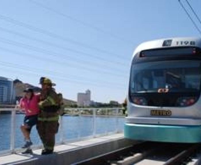 A firefighter assists a person off a metro light rail train near a waterfront on a sunny day.