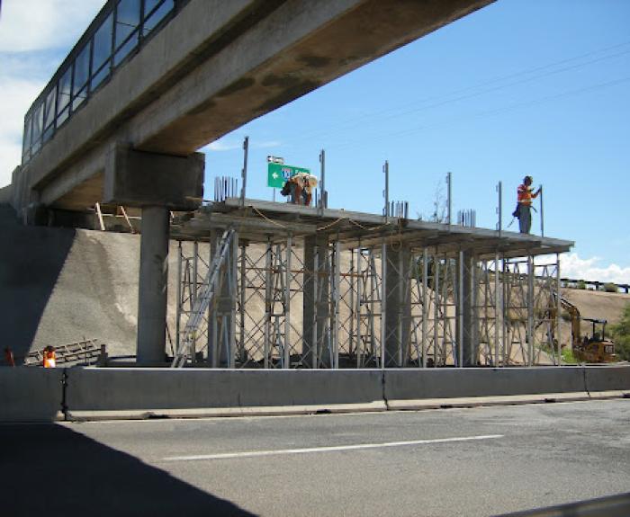 Two construction workers stand on scaffolding under an overpass, working on a concrete structure above a road with vehicles and equipment nearby.