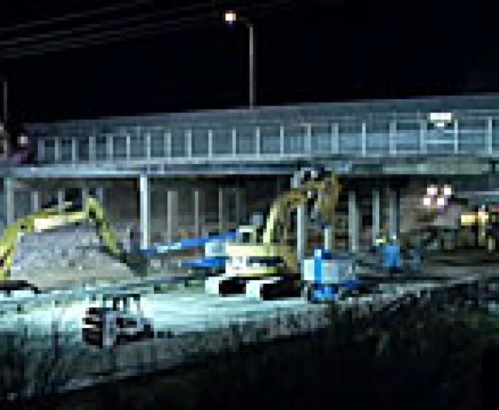 Construction equipment and vehicles work under a highway overpass at night, illuminated by overhead lights.