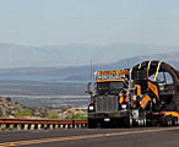 A large black semi-truck marked Oversize Load transports heavy equipment on a flatbed trailer along a highway with mountains in the distance.