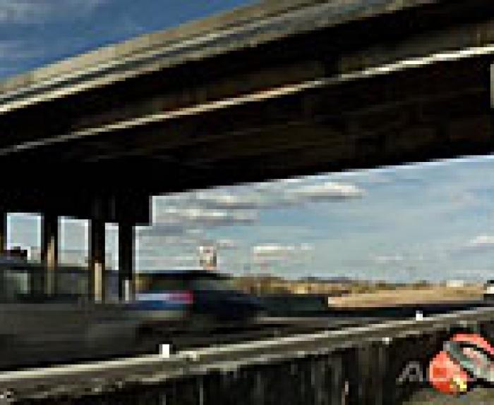 Cars driving quickly under a highway overpass, with fields in the background.