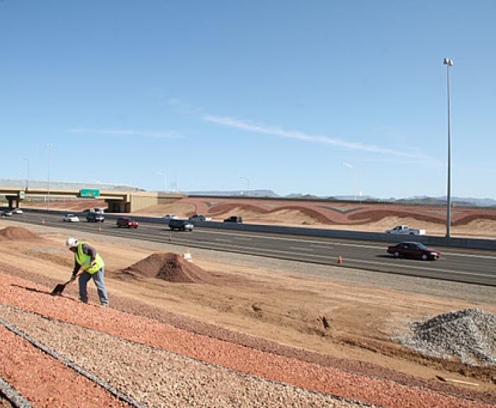 A construction worker in a yellow vest works on a dirt embankment beside a busy multi-lane highway under a clear blue sky.