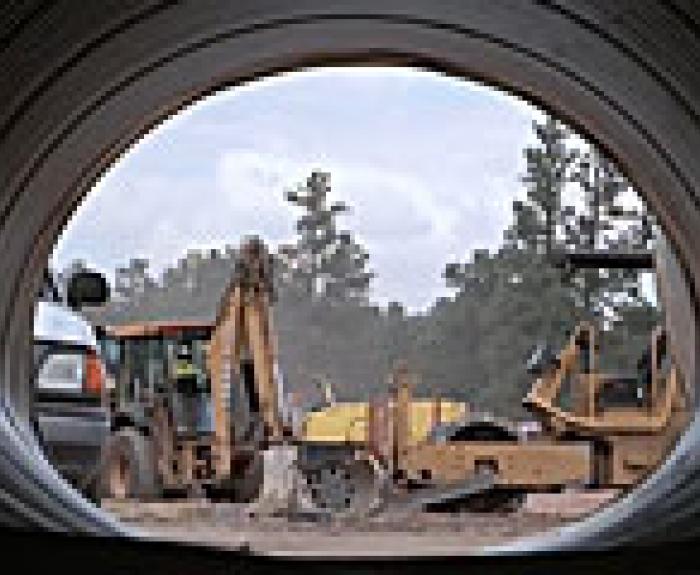 View from inside a large pipe showing construction vehicles, including a backhoe and bulldozer, working at a construction site with trees in the background.