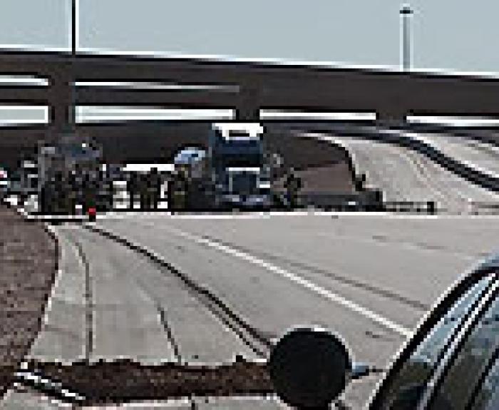 Police vehicles and a semi-truck block a highway on-ramp under an overpass, with officers and emergency personnel standing nearby.