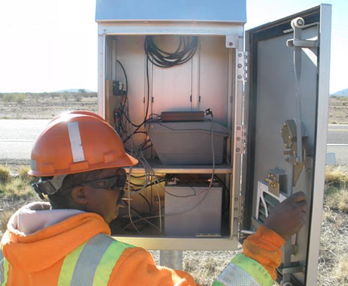 A person wearing an orange safety helmet and reflective jacket inspects the open control panel of a roadside electronic equipment box.