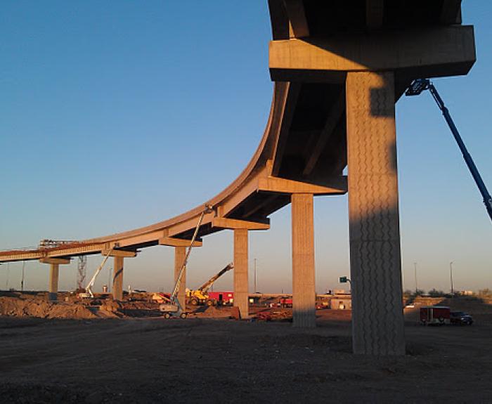 A large concrete highway overpass under construction, supported by tall pillars, with cranes and construction equipment on the dirt ground below.