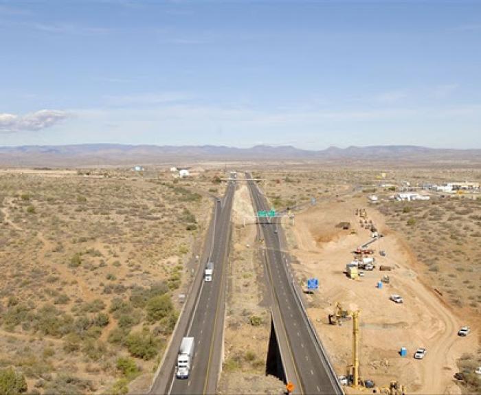 A highway stretches through a dry landscape with cars and construction equipment on the roadside.