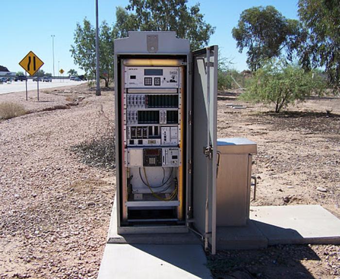 An open roadside utility cabinet reveals electronic equipment, wires, and devices inside, situated on a concrete pad near a road in a dry, desert area.