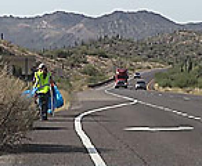 A person in a yellow vest walks along the shoulder of a highway, carrying a large blue bag, with cars and trucks on the road and mountains in the background.