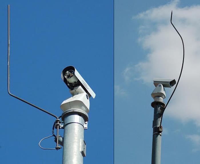 Two surveillance cameras are mounted on metal poles. The left cameras antenna is straight, while the right cameras antenna is bent upwards and twisted.