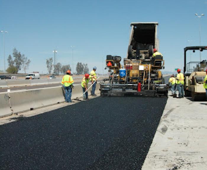 Construction workers pave a highway using large machinery. Fresh asphalt is being laid, and workers in safety vests are standing on both sides of the road.